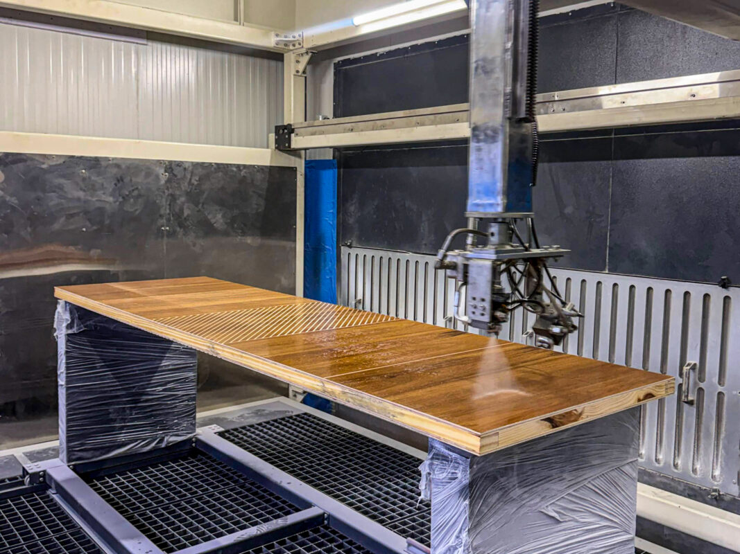 Robotic spray arm applying an even PU finish to a grooved teak veneer door panel inside the LK Doors and Interiors dust-free spray booth